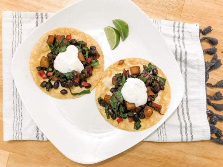 gluten free vegetarian tostadas with corn tortillas black beans swiss chard and potato topped with honey lime yogurt sauce on white plate with lime wedges on tea towel horizontal image
