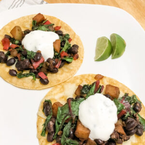 gluten free vegetarian tostadas with corn tortillas black beans swiss chard and potato topped with honey lime yogurt sauce on white plate with lime wedges vertical image