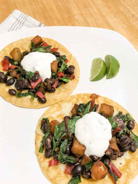 gluten free vegetarian tostadas with corn tortillas black beans swiss chard and potato topped with honey lime yogurt sauce on white plate with lime wedges vertical image