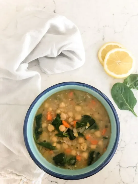 bowl of greek lemon orzo soup on counter with teatowel, spinach, and lemon slices