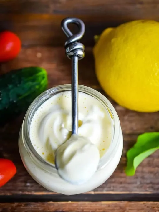 Jar of homemade gluten free mayo with spoon on wooden table with lemon, cucumber, and tomato