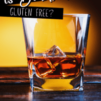 Square glass with bourbon on the rocks sitting on a dark countertop against a yellow shadowed wall and text overlay that says is bourbon gluten free?