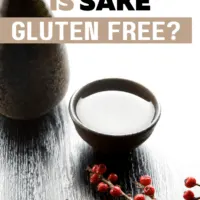 Sitting on a wooden table top is a black porcelain bottle with a matching porcelain cup filled with sake sitting beside a branch with red berries for decoration and a text overlay that reads Is Sake Gluten free