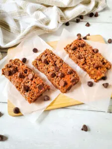 3 chocolate nut free gluten free protein bars on parchment with chocolate chips scattered on wooden board with tea towel