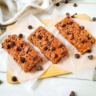 3 chocolate nut free gluten free protein bars on parchment with chocolate chips scattered on wooden board with tea towel