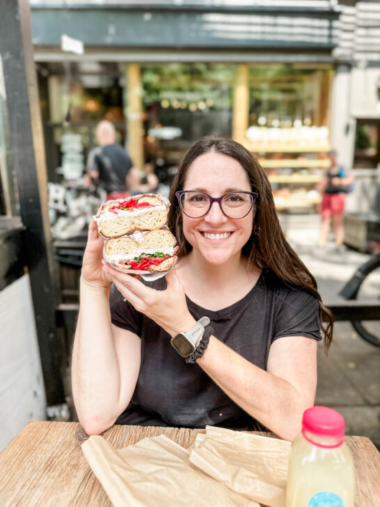 Sharon McCaskill from The Helpful GF holding up a gluten free bagel with vegan cream cheese and veggies in front of modern bread and bagel in NYC