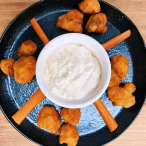 crispy baked cauliflower bites on a black plate with carrot sticks and bowl of dip in the middle