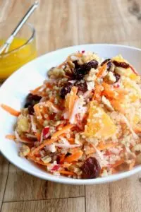 orange cranberry quinoa salad with carrots on white bowl on top of a wooden table with dressing in jar