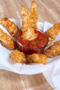 quinoa crusted chicken fingers on white paper dipped in salsa