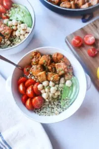 pesto chicken quinoa bowl with avocado, chickpeas, and tomato in white bowl with tomatoes on cutting board