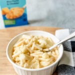 high protein gluten free mac and cheese in a white bowl with spoon on top of a wooden board with a checked tea towel and a box of annie's gluten free mac and cheese in background