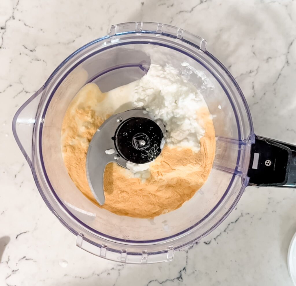 a blender sitting on a counter top that has cauliflower, cheese powder, milk, and cottage cheese inside of it.