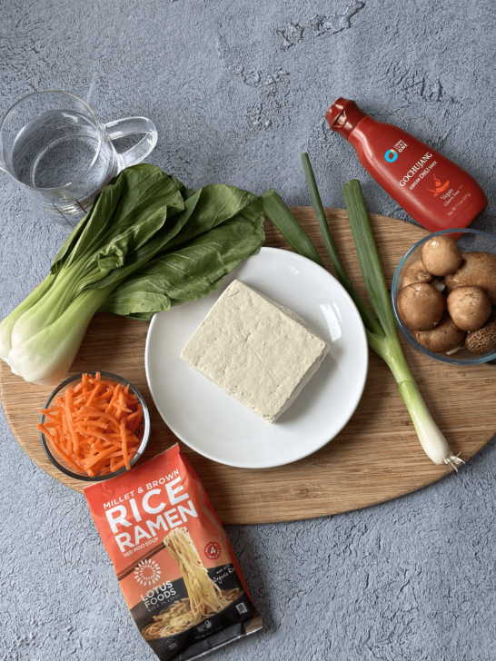 Ingredients for simple gluten-free ramen recipe white dish of tofu, measuring cup of water, glass bowl of shredded carrots, package of rice ramen, green onion, glass bowl of mushrooms, and bottle of gochujang