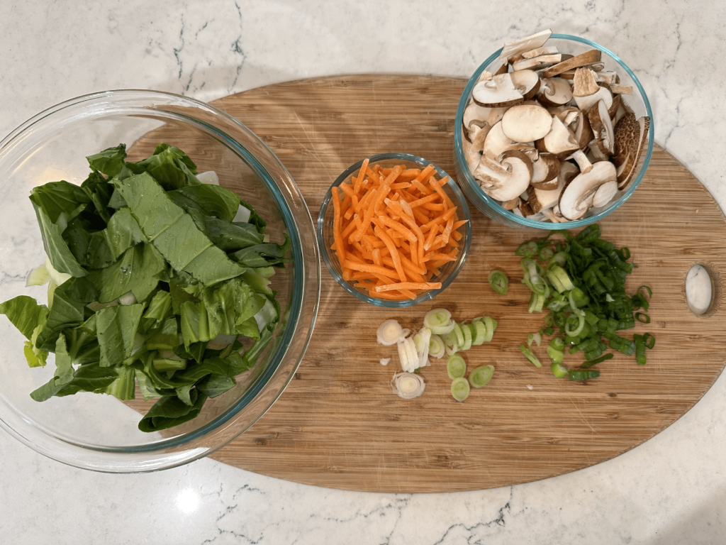 Vegetable ingredients sitting on wooden cutting board, glass bowl of baby bok choy, small glass bowl of shredded carrots, glass bowl of sliced mushrooms, chopped up green onions