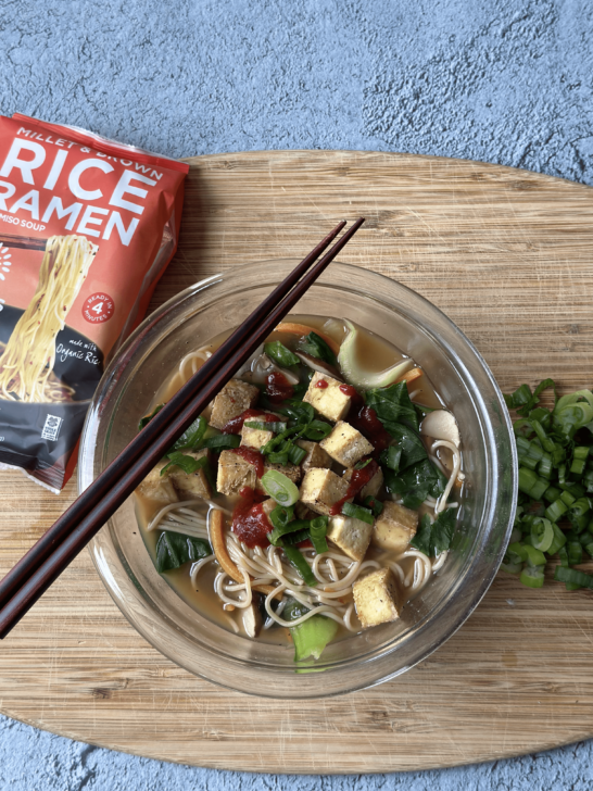 gluten free ramen soup in a clear bowl with chopsticks on top of a wooden board with a package of rice ramen in on the left side and chopped up green onions on right side