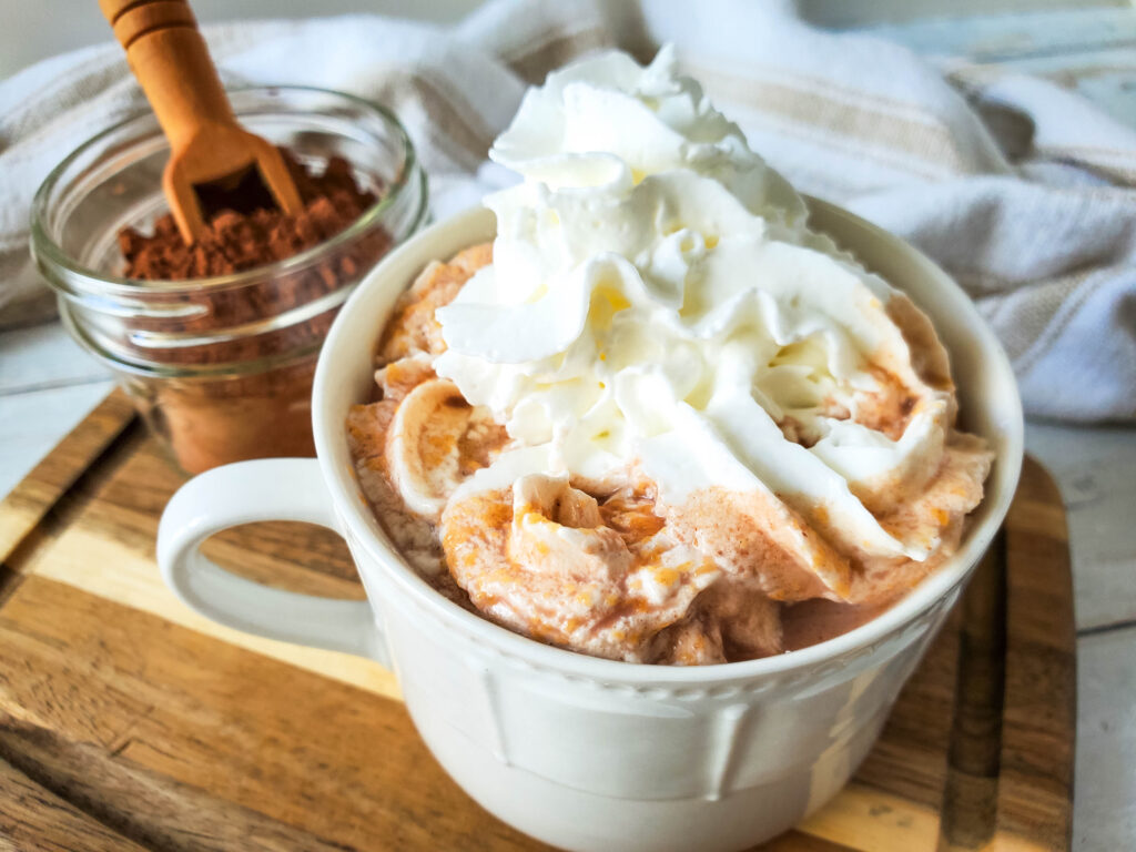 A white mug with peanut butter hot chocolate that's topped with whipped cream is sitting on a cutting board next to a jar of cocoa powder.