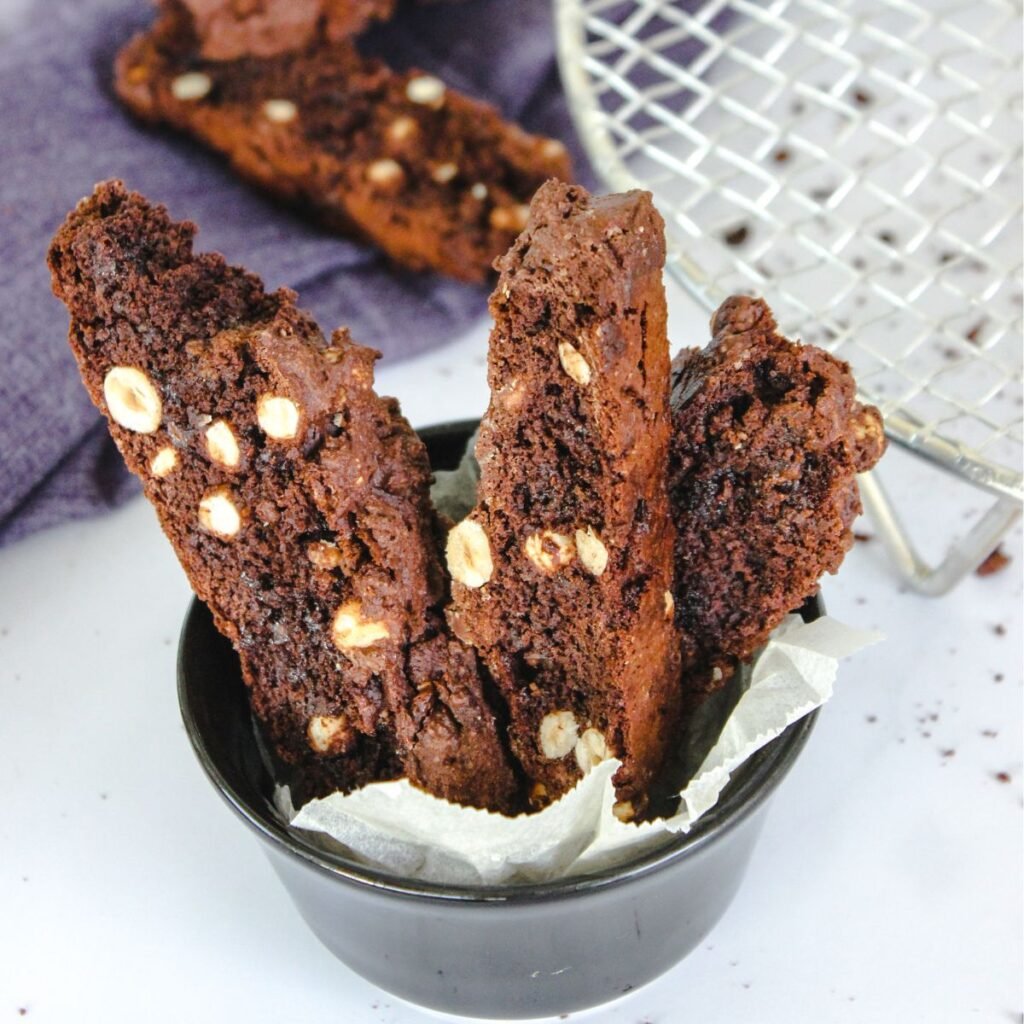 thumbnail image of 3 pieces of gluten free chocolate hazelnut biscotti in a black cup with some in a background on a blue tea towel