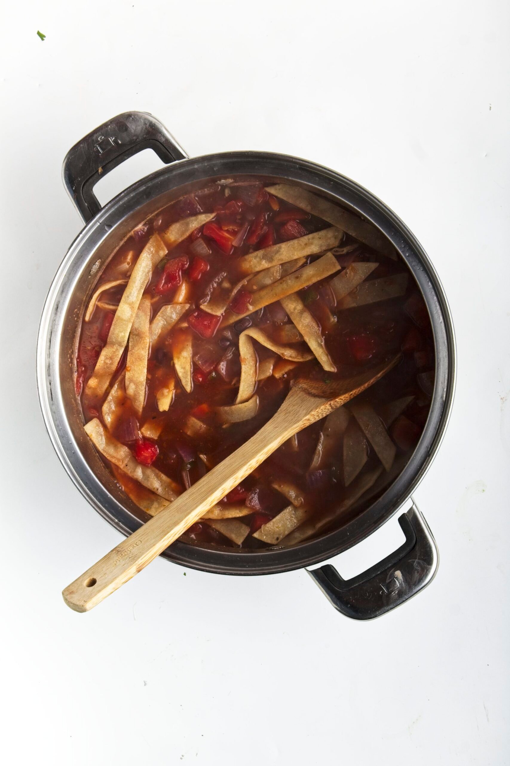 A soup pot cooking tortilla soup with a wooden spoon and crispy tortilla strips