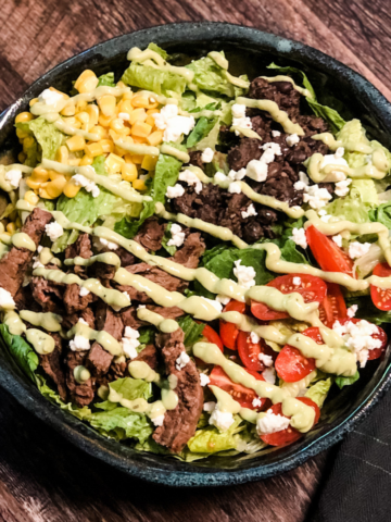 gf salad topped with steak, corn, black beans, tomatoes, and avocado dressing in a pottery bowl on a wooden table with black tea towel