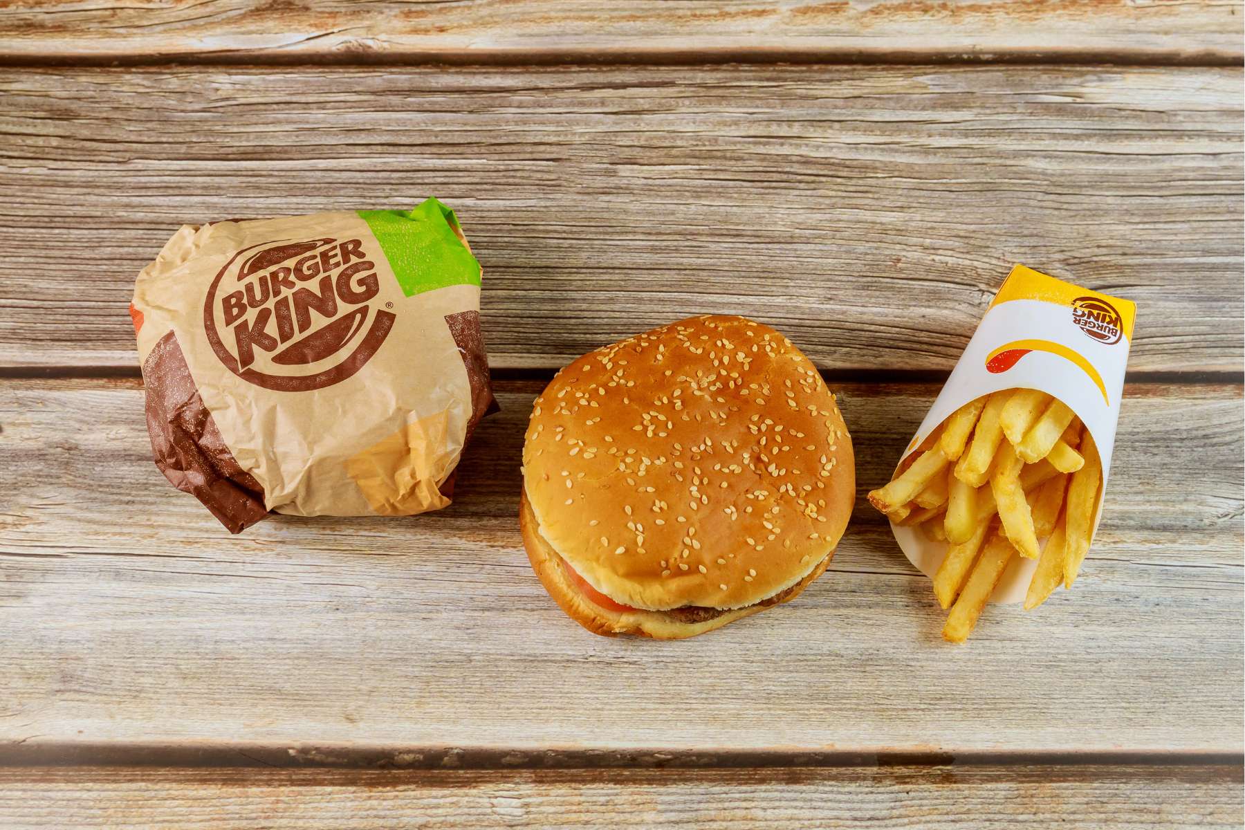 An array of Burger King foods including a wrapped burger, an unwrapped burger and container of fries are sitting on a wooden table.