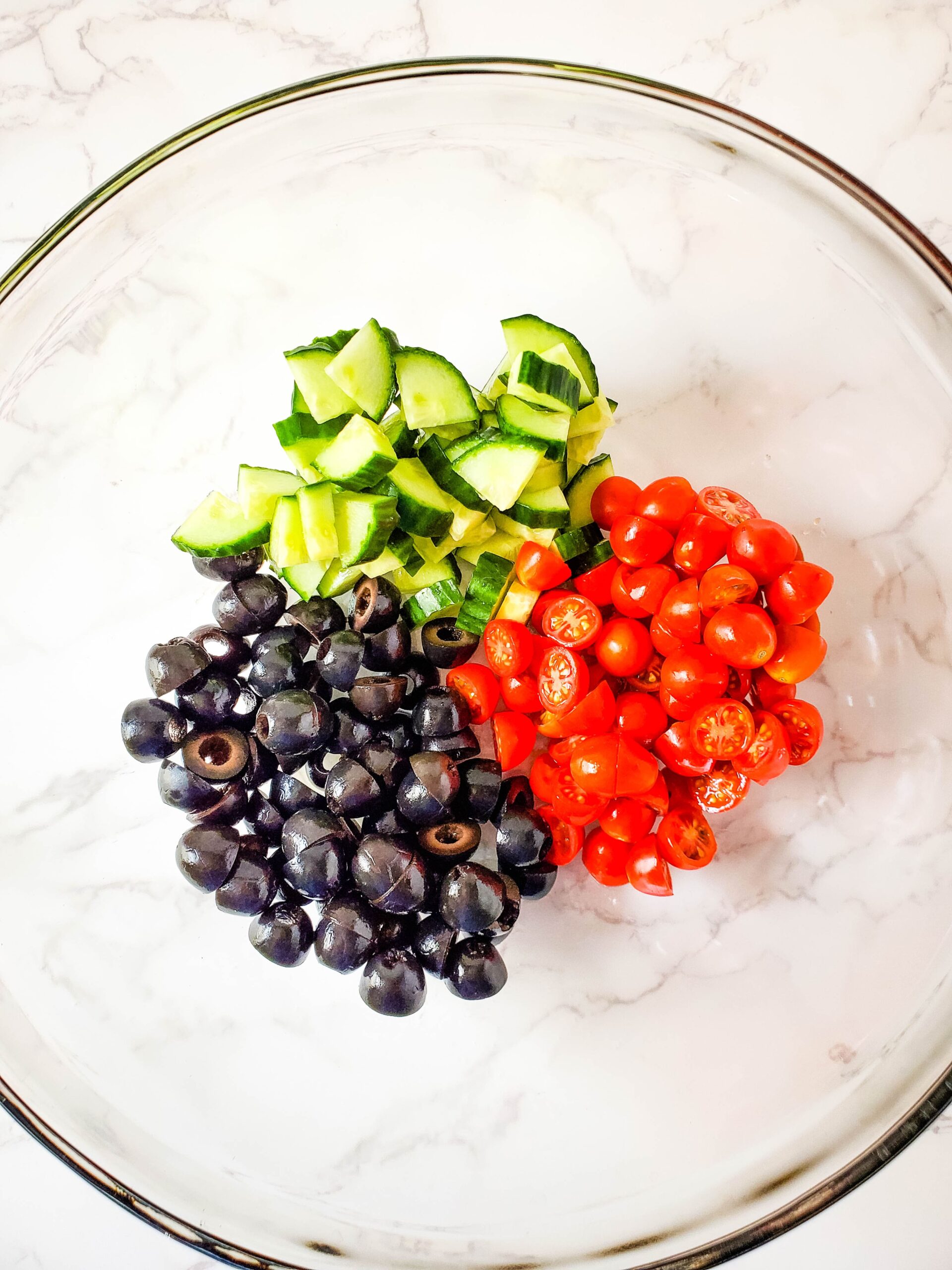 A glass bowl sitting on a white marbled countertop is filled with sliced cucumbers, tomatoes and black olives