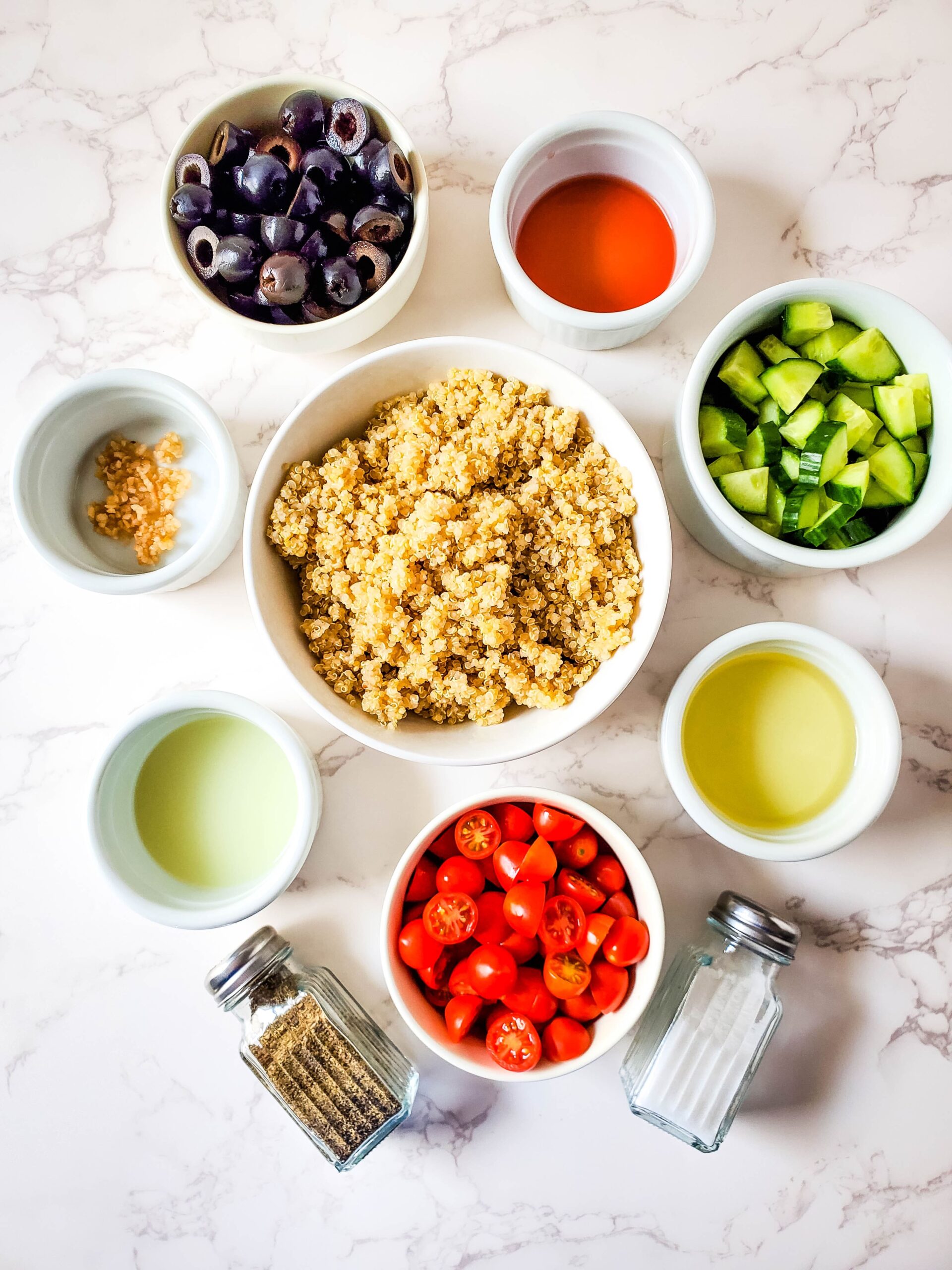 On a white marbled countertop are several white bowls. In the middle is a large bowl of cooked quinoa. In smaller bowls surrounding the large bowl are black olives, red wine vinegar, cucumber, oil, tomatoes, lemon juice, and garlic. Underneath the bowls are salt and pepper shakers.