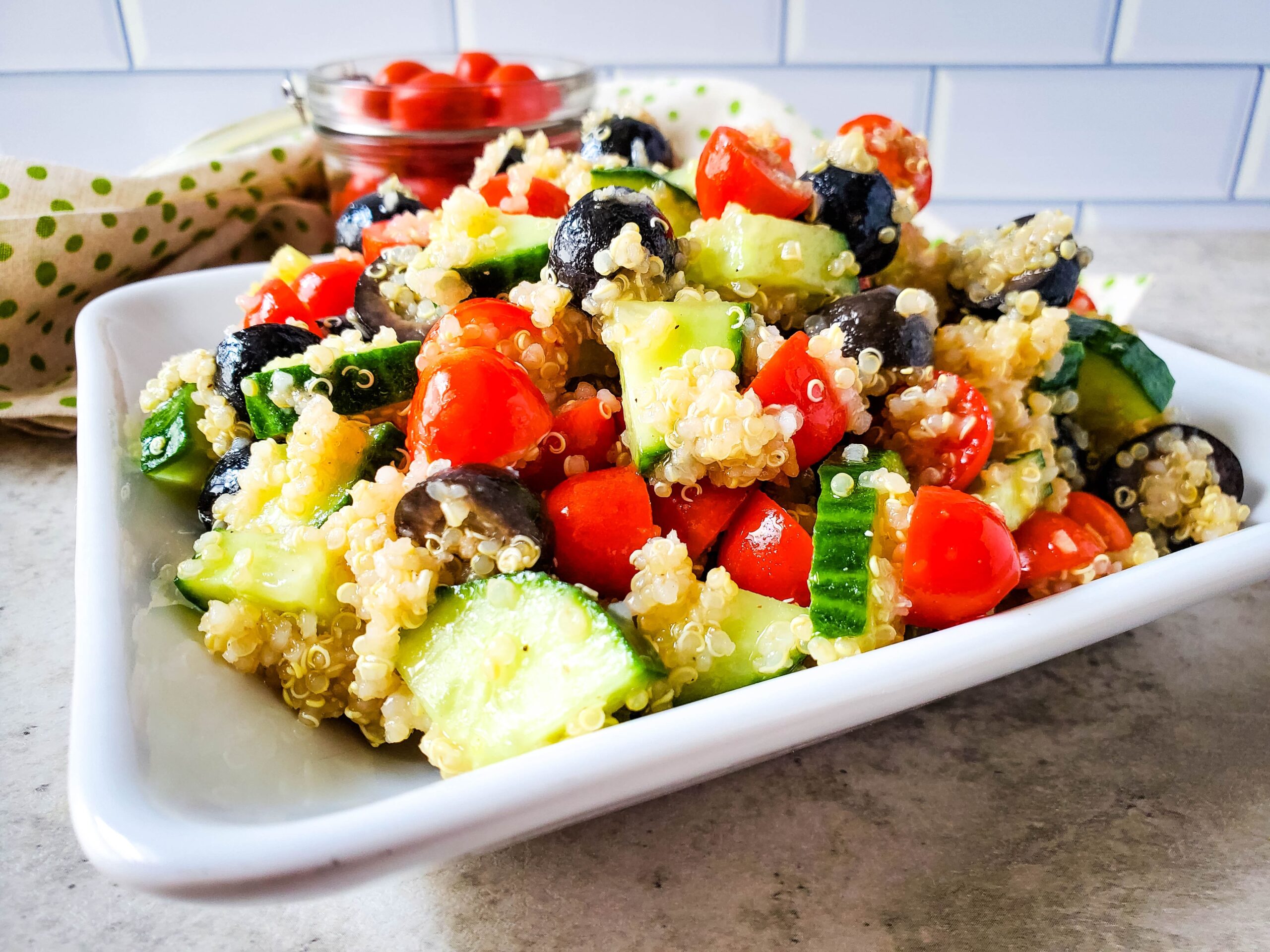 A square white plate filled with Mediterranean Quinoa Salad is sitting on a counter top.