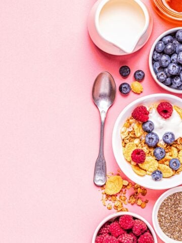 A pink background and down the right-hand side is a variety of high fiber items including cereal, blueberries, raspberries and a bowl of seeds. There is also a small pitcher of milk and a spoon.
