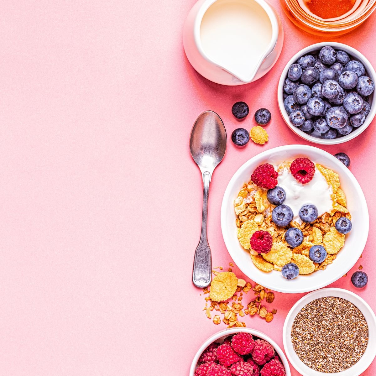 A pink background and down the right-hand side is a variety of high fiber items including cereal, blueberries, raspberries and a bowl of seeds. There is also a small pitcher of milk and a spoon.