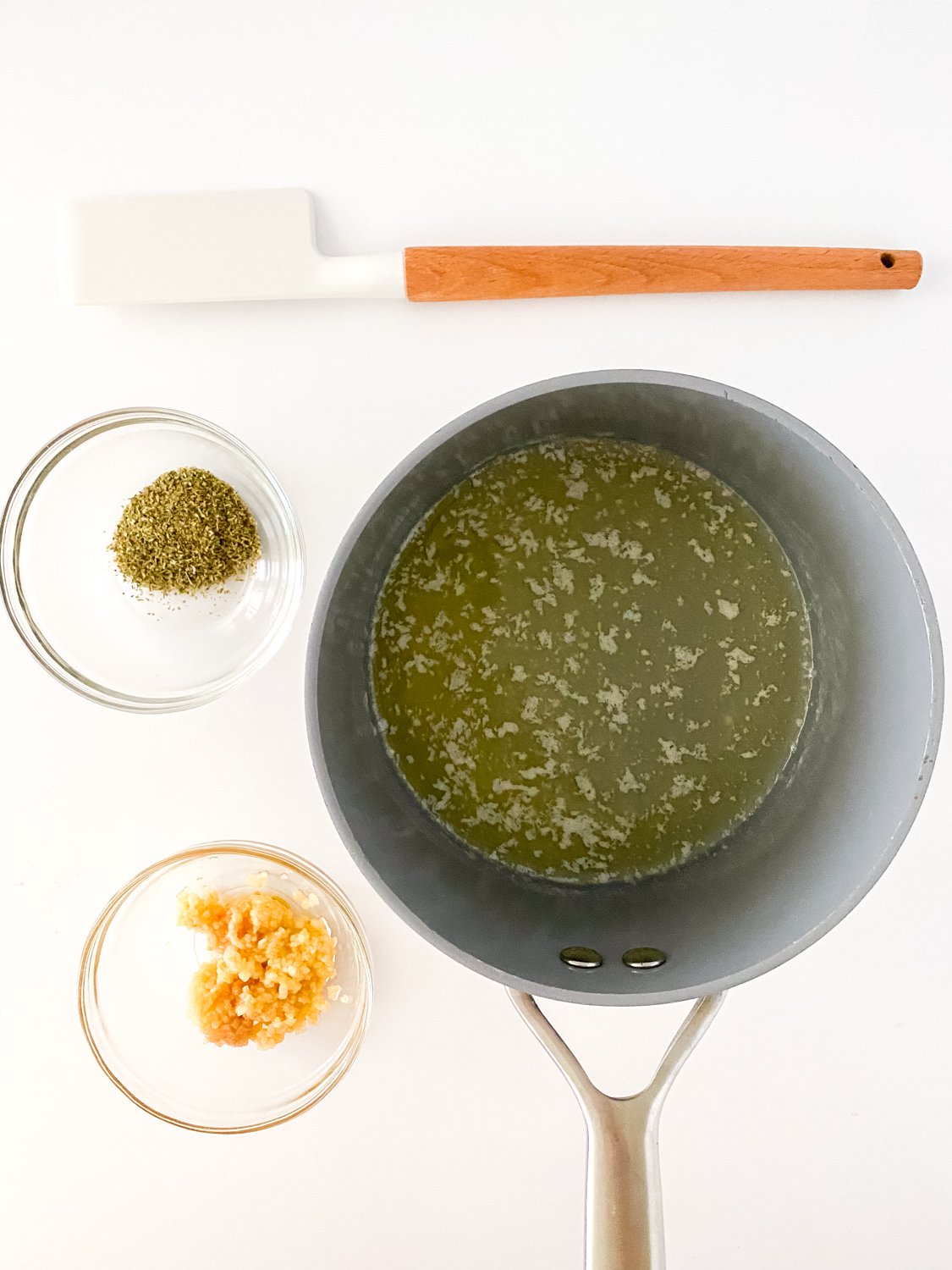 On a counter is a rubber spatula that is sitting above a pot with melted butter and 2 small glass bowls with ingredients to make cream garlic parmesan sauce