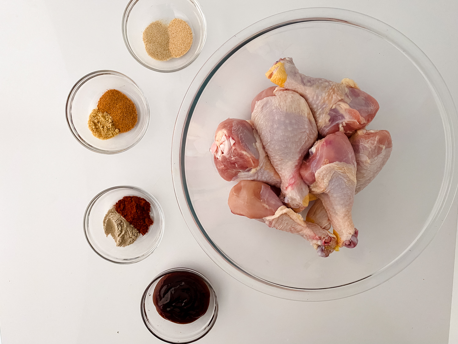 A large glass bowl with raw chicken drumsticks is surrounded by small glass bowls filled with ingredients for BBQ Chicken Drumsticks