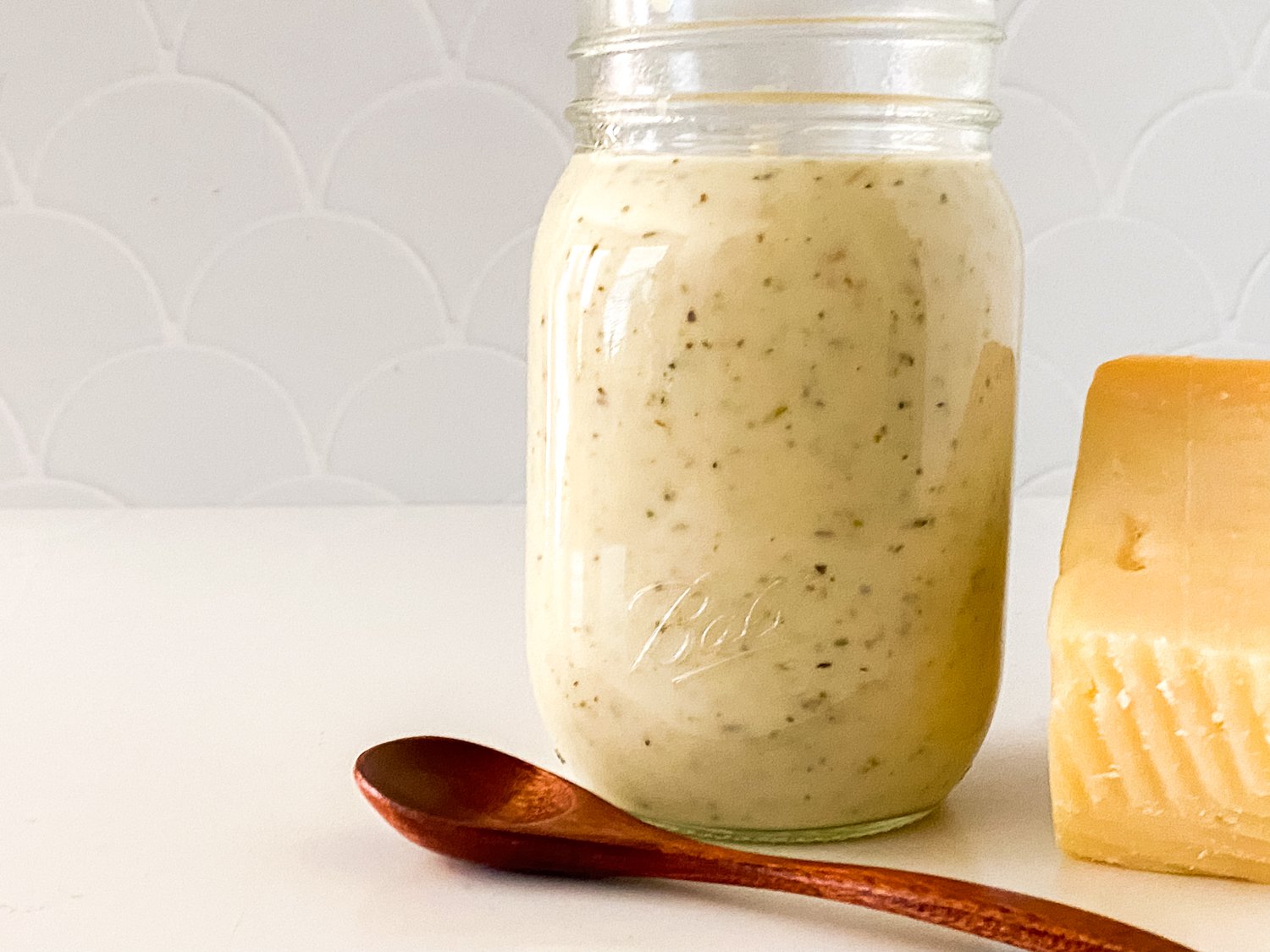 A jar of fresh, homemade creamy garlic parmesan sauce sitting beside a block of parmesan cheese and a wooden spoon