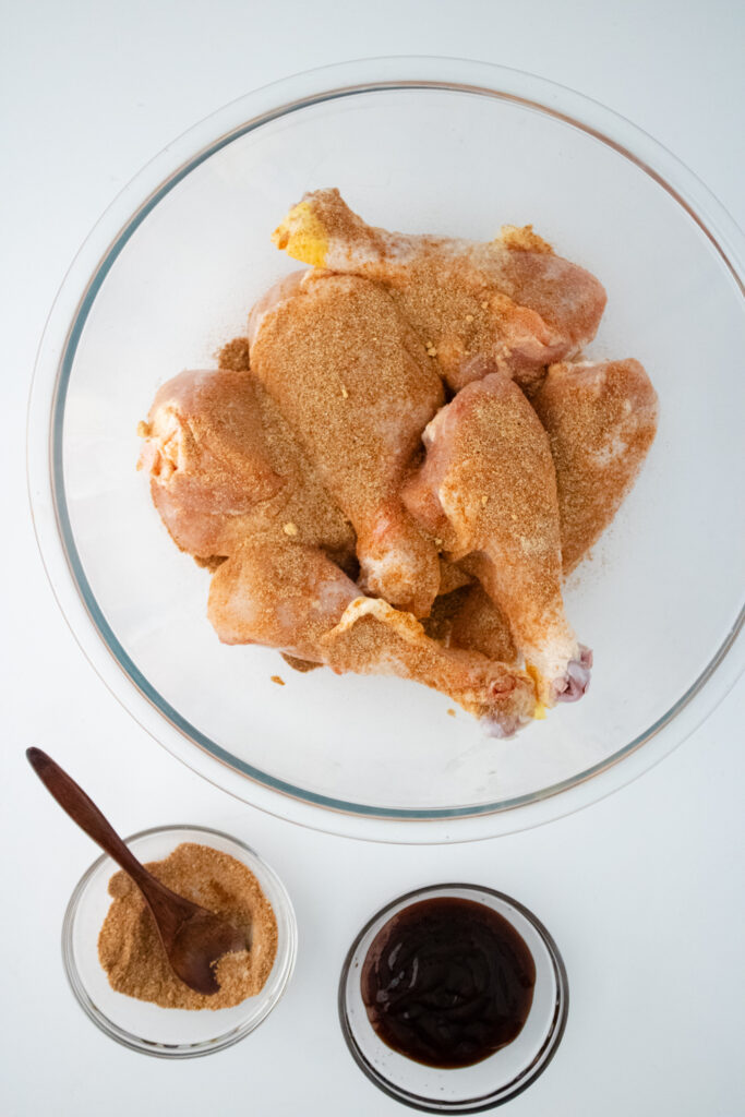 A glass bowl containing raw chicken legs that are covered in spices