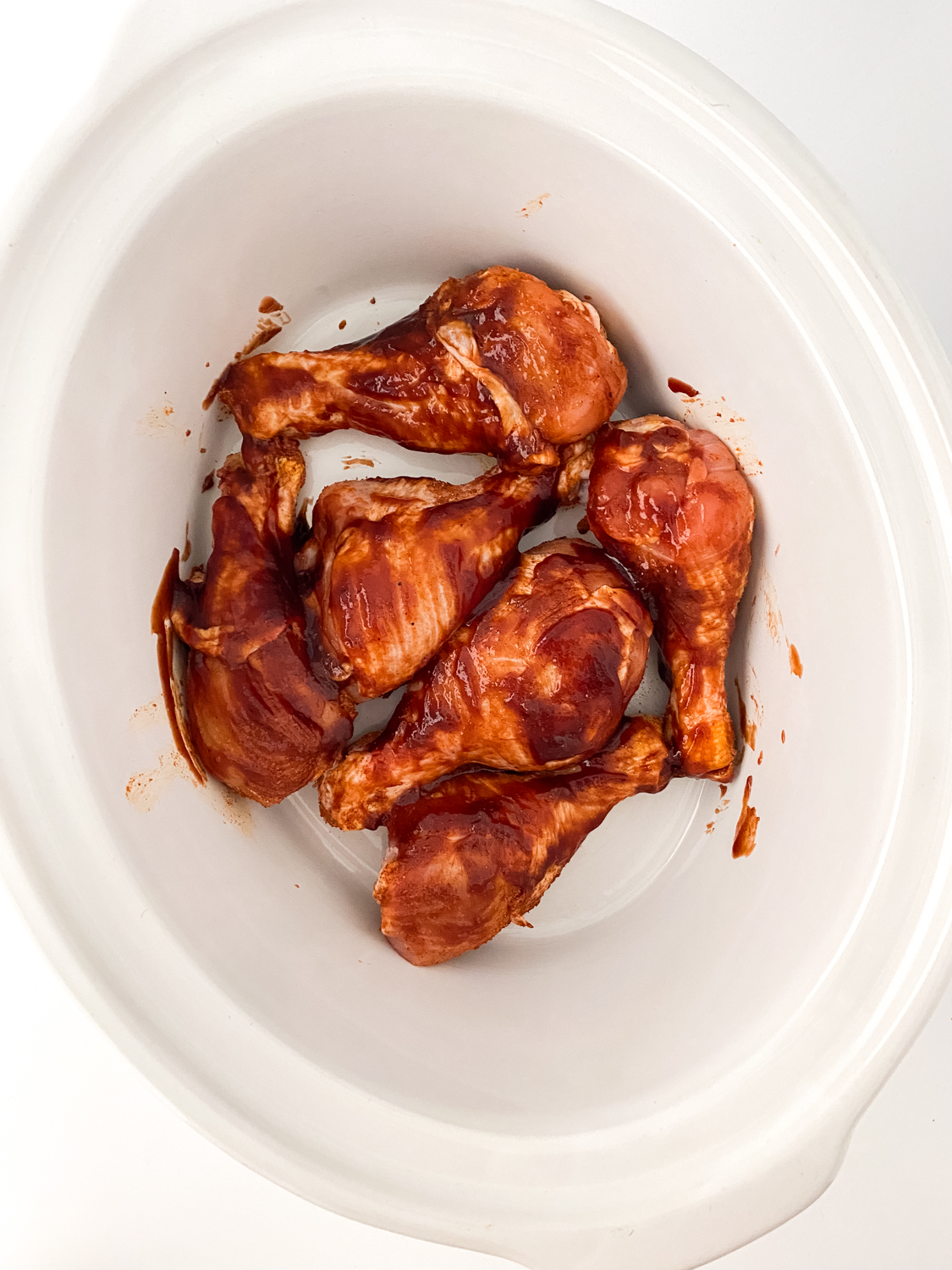 Chicken legs covered in BBQ sauce in the bottom of a white slow cooker