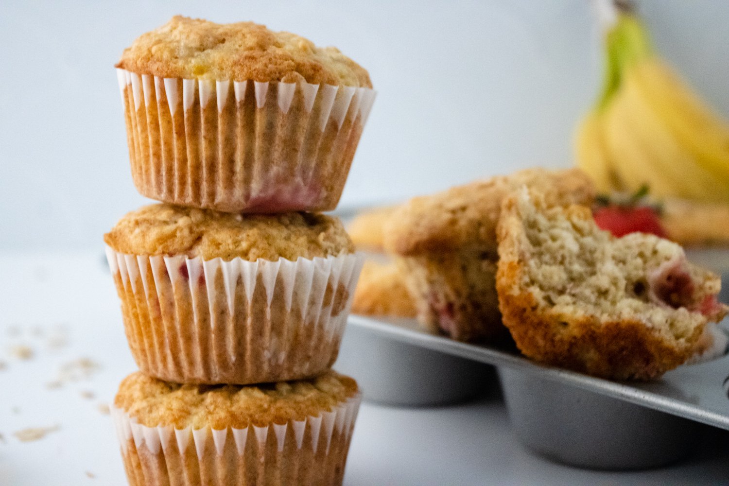 3 gluten-free strawberry banana muffins are stacked on top of each other. In the background is a muffin tin with muffins in it and a bunch of bananas.