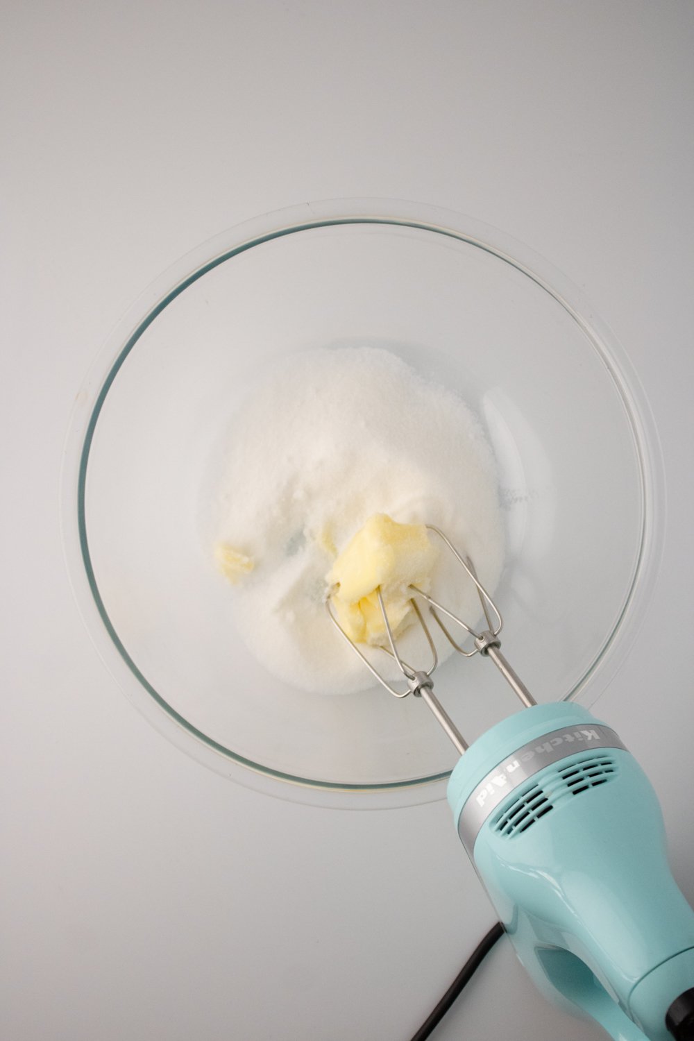 Butter and sugar have been added to a glass bowl. A light blue hand mixer is resting on the bowl.