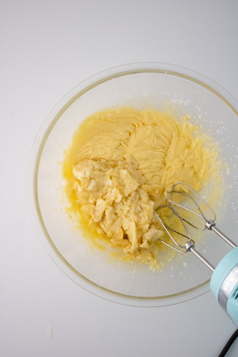 A glass bowl has creamed butter and sugar, and mashed bananas have been added in. A light blue hand mixer is resting on the bowl.