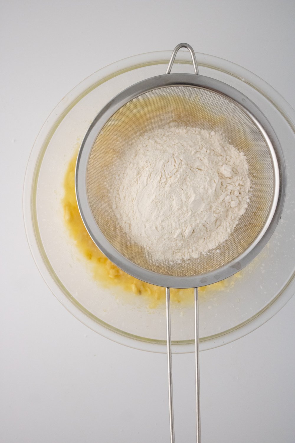 A metal sieve is filled with flour and resting on top of a glass bowl with the wet ingredients for strawberry banana muffins.