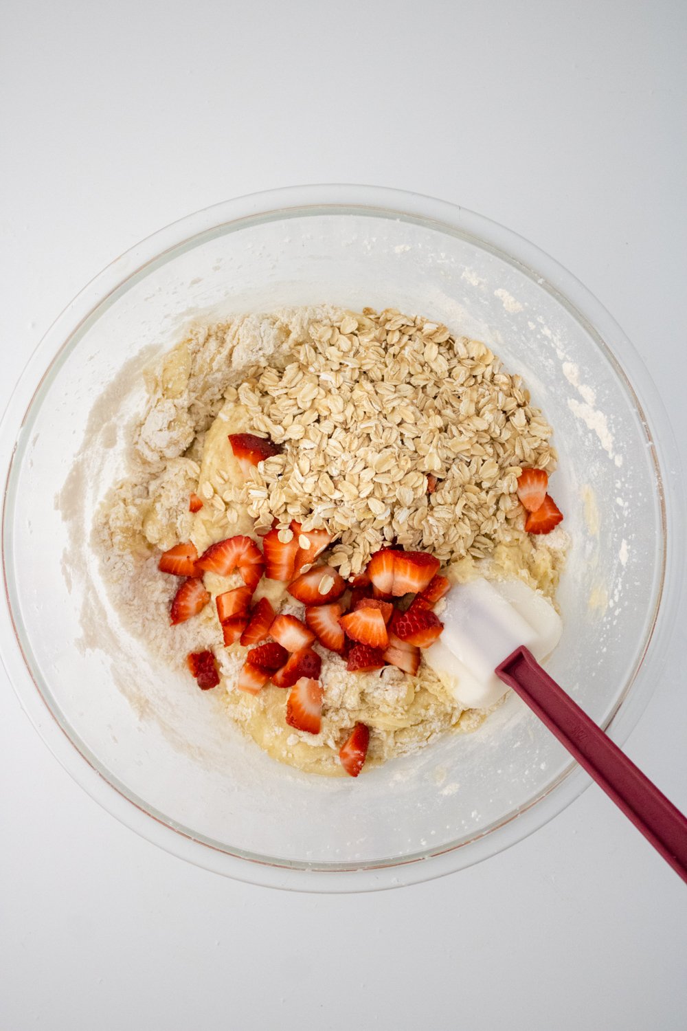 Oats and diced strawberries have been added to a glass bowl with the mixed ingredients for healthy strawberry banana muffins. A rubber spatula with a red handle is resting inside the bowl.