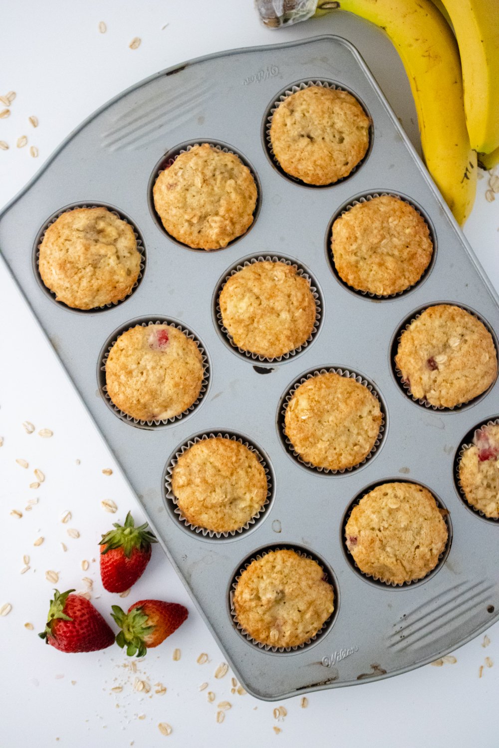 A top-down view of a muffin tin with freshly baked Gluten Free Strawberry Banana Muffins. A bunch of bananas is sitting on the right side of the tin and three fresh strawberries are sitting on the left side.