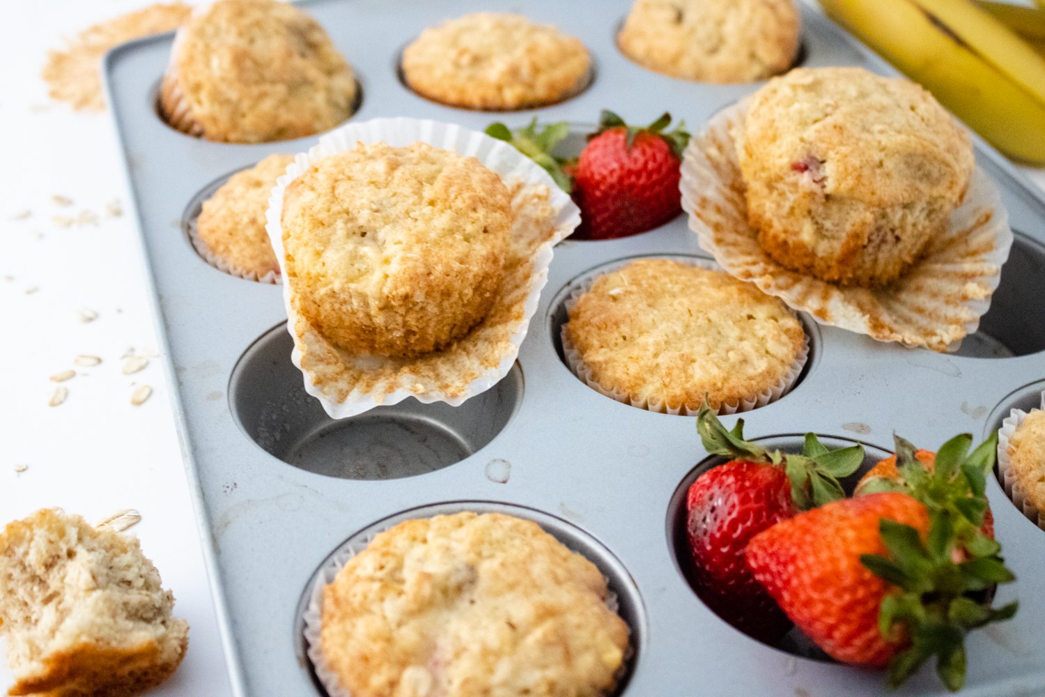 A close-up of a muffin pan. Some muffins are in the tin, some are sitting on top of the tin, and 2 cups have fresh strawberries in them.