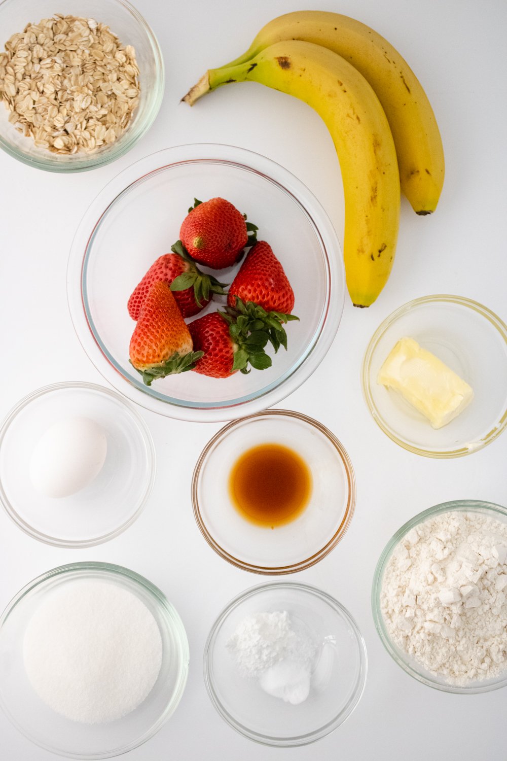 The ingredients for Gluten Free Strawberry Banana Muffins are sitting in glass bowls on a white countertop.