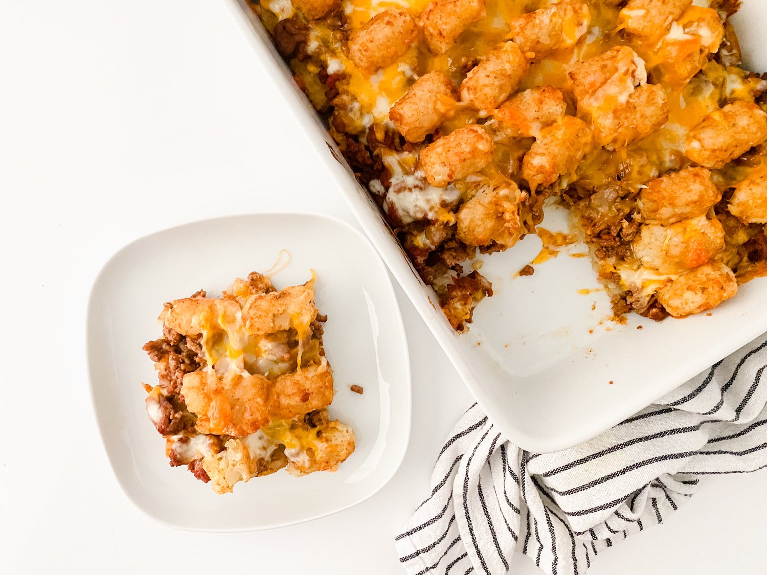 A piece of tater tot casserole has been taken out of a white casserole dish and placed on a white plate beside it. The casserole dish is sitting on a black and white striped tea towel.