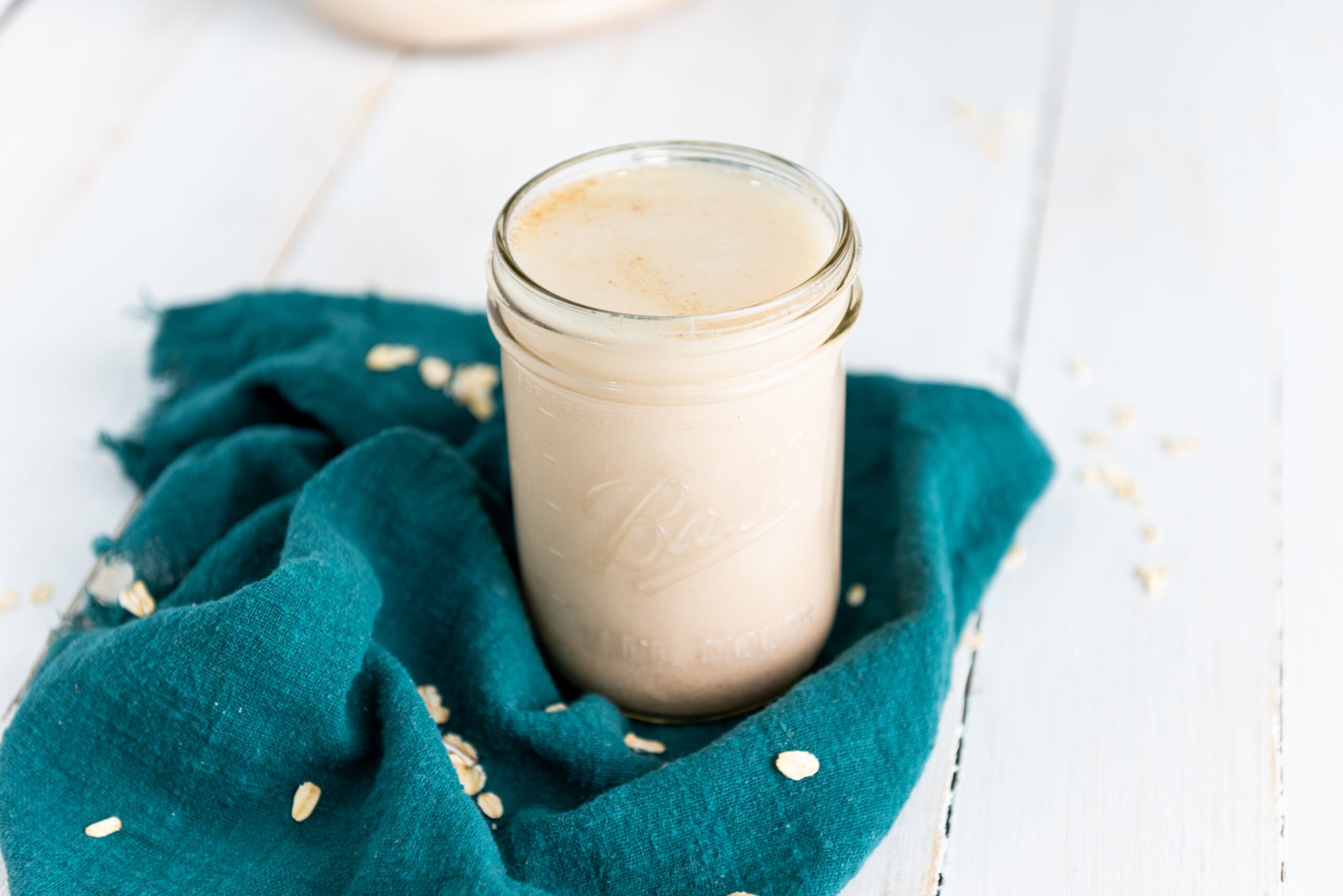 A small mason jar of homemade oat milk sits on top of a teal colored cloth with oats sprinkled around it