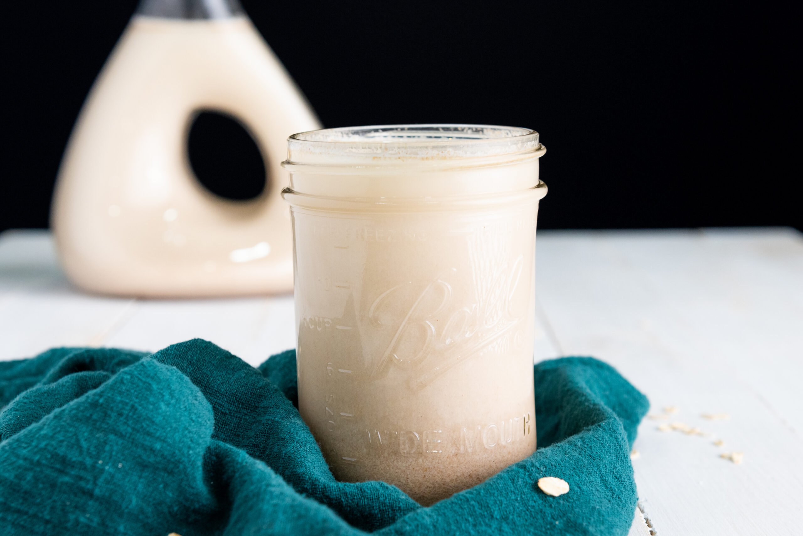 In the foreground is a glass jar filled with gluten free oat milk. In the background a glass decanter is also filled with oatmeal milk