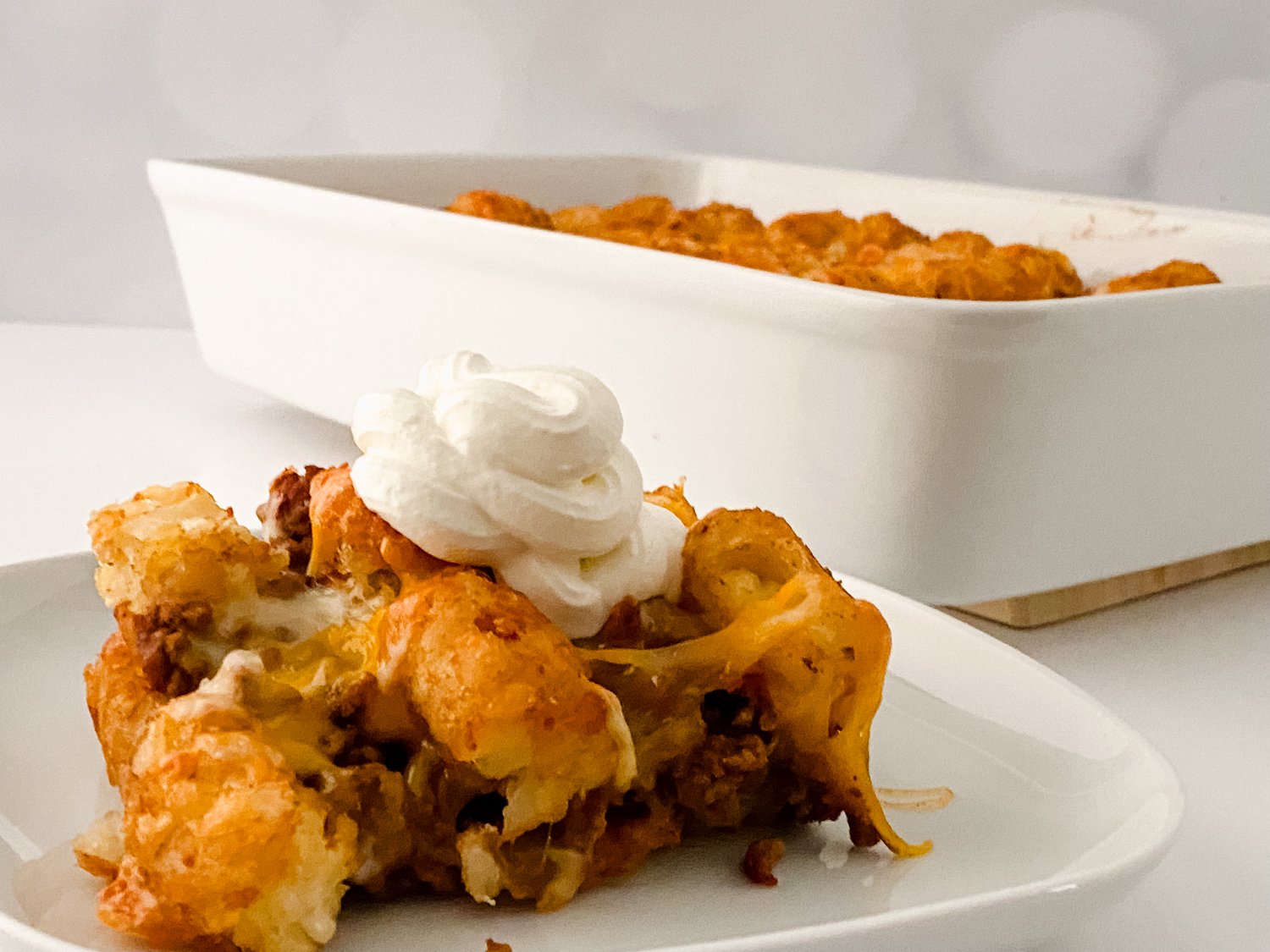 In the foreground is a plate with a square piece of tater tot casserole topped with sour cream. In the background is a white casserole dish with taco tot casserole.