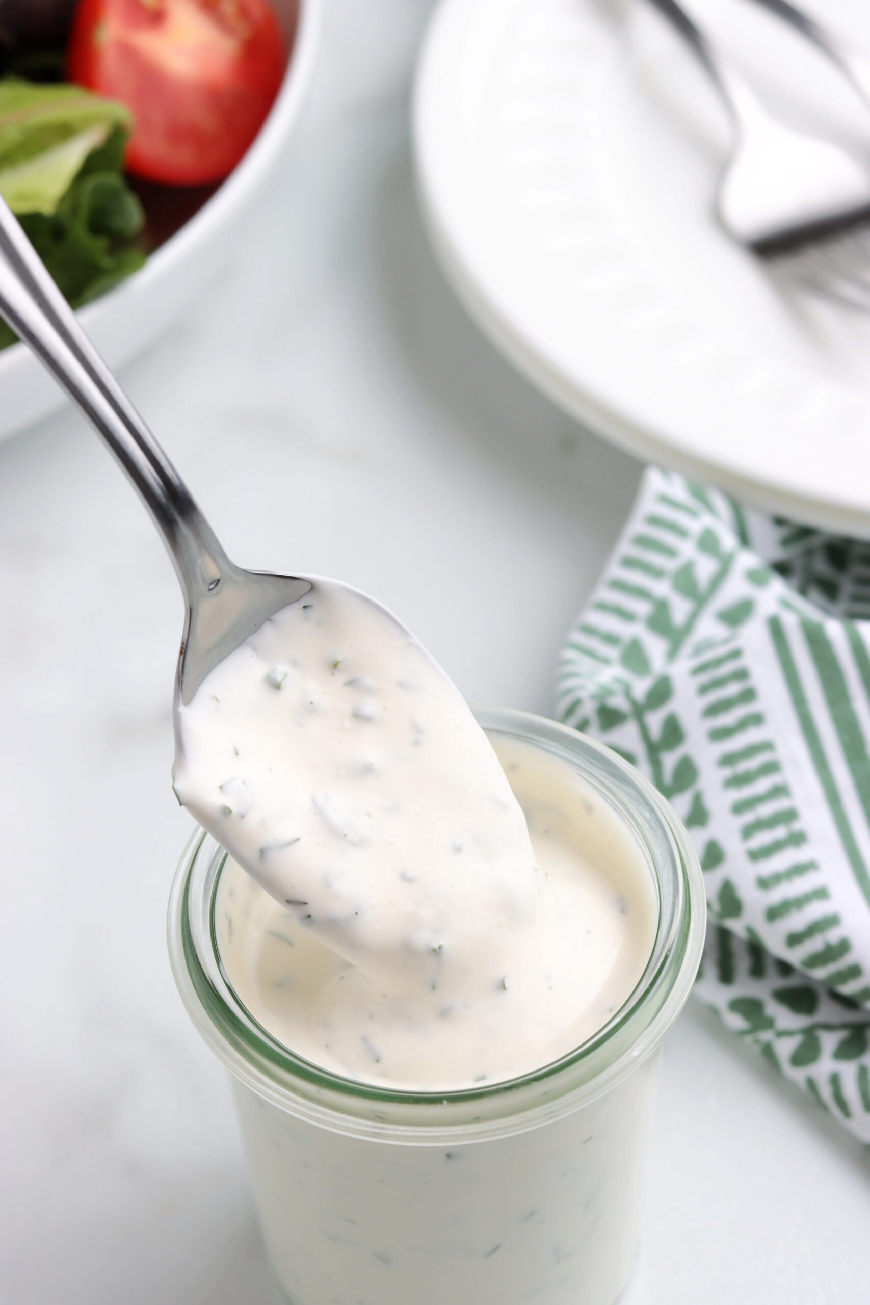 A spoon is scooping ranch dressing from a glass jar. The background is blurred and has a side salad next to a white salad plate with a fork.