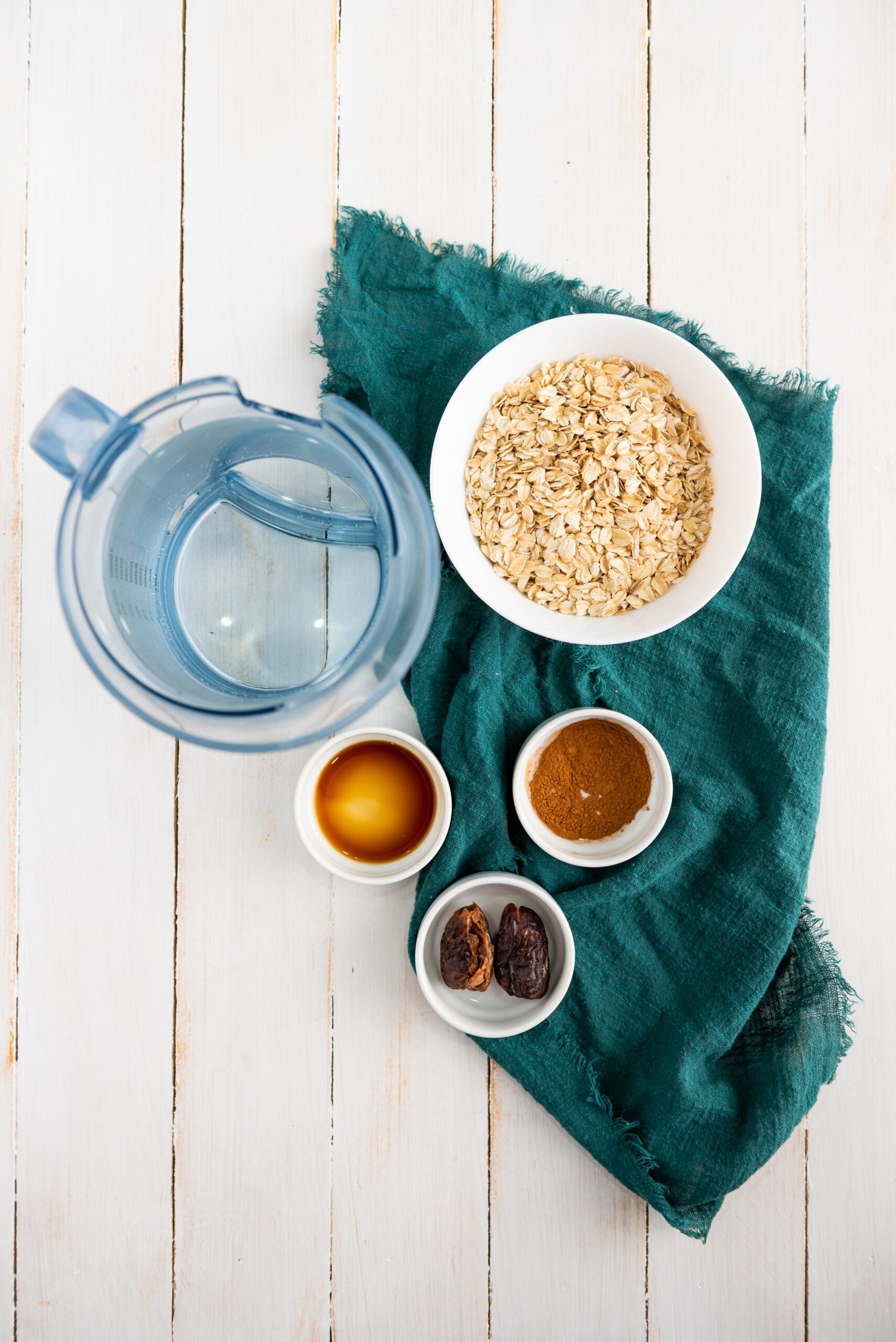 The ingredients to make gluten-free oat milk sit on a white, wooden countertop. Underneath the white bowls containing the ingredients, there is also a dark green cloth placemat.