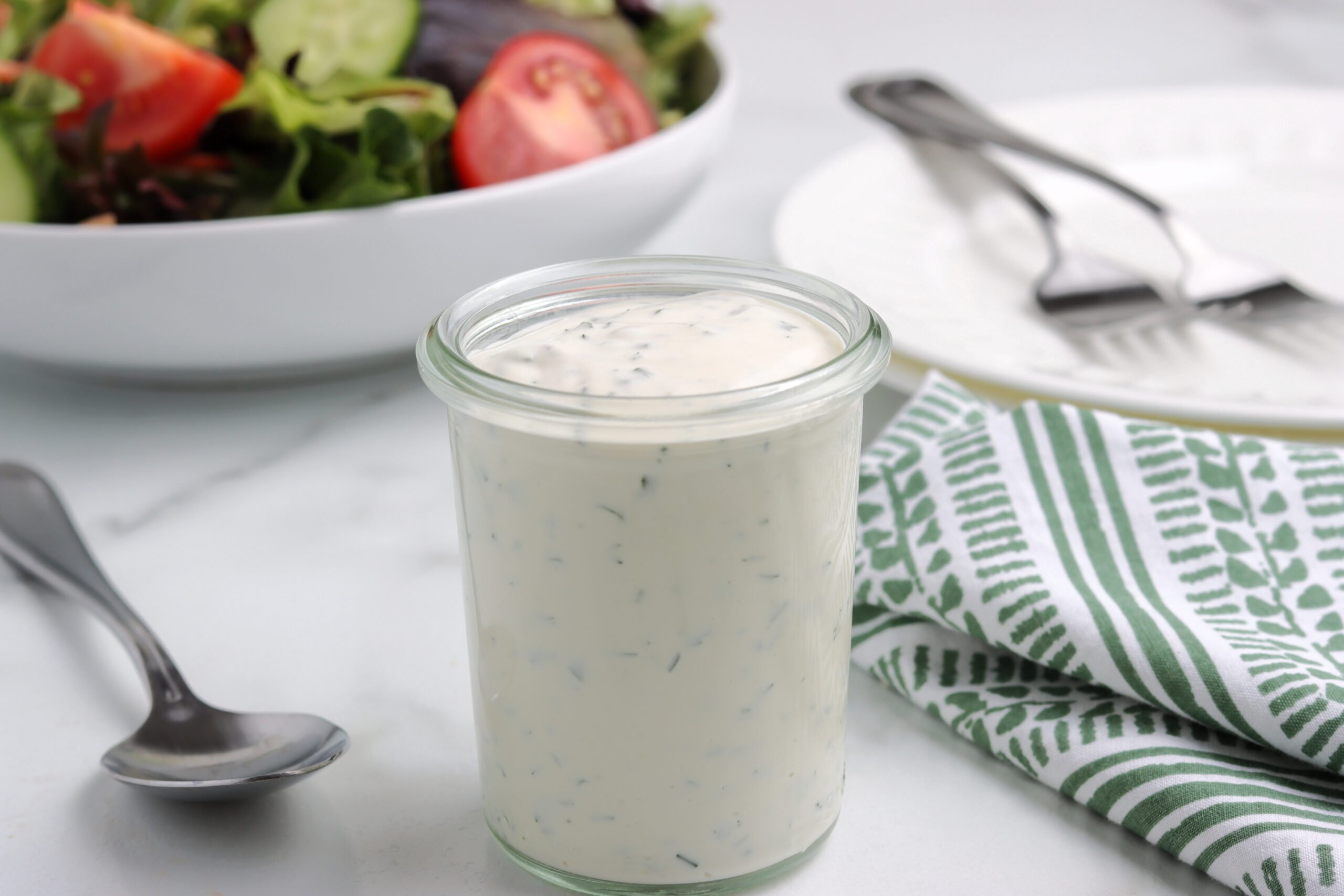 A small glass jar of homemade gluten-free ranch sits in the foreground. In the background is a salad in a white bowl beside a white salad plate.