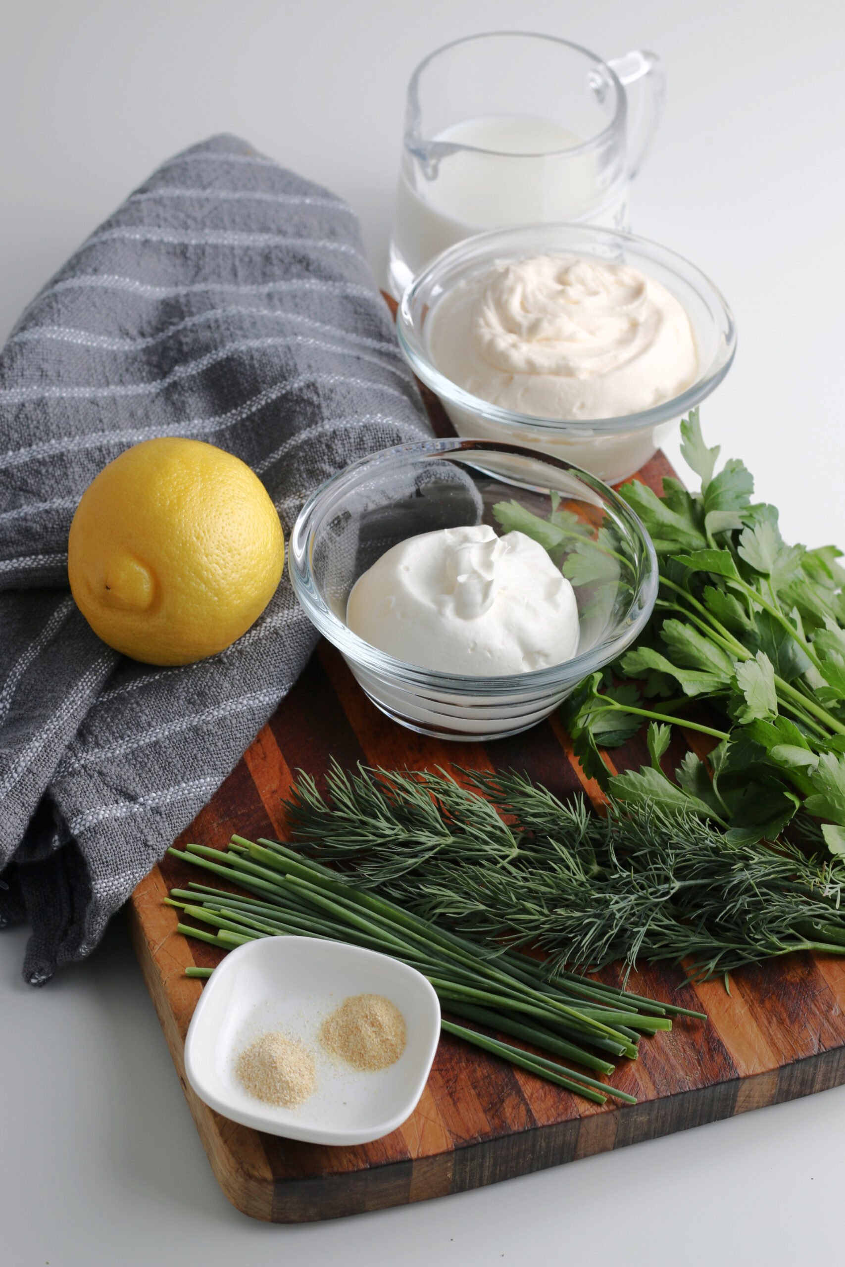All of the ingredients for homemade ranch dressing sit on a wooden cutting board with a grey tea towel.